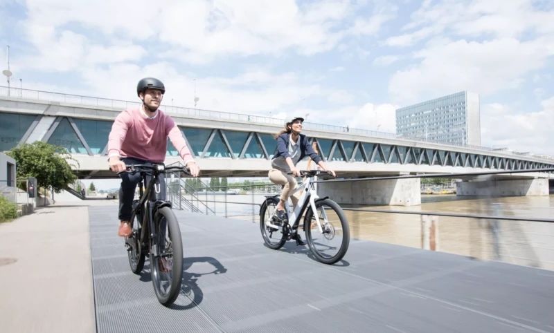 Eine Frau und ein Mann, beide mit Fahrradhelmen, unterhalten sich lächelnd. Die Frau steht neben einem E-Bike, während der Mann auf einer Bank sitzt, neben ihm ein Rennrad.