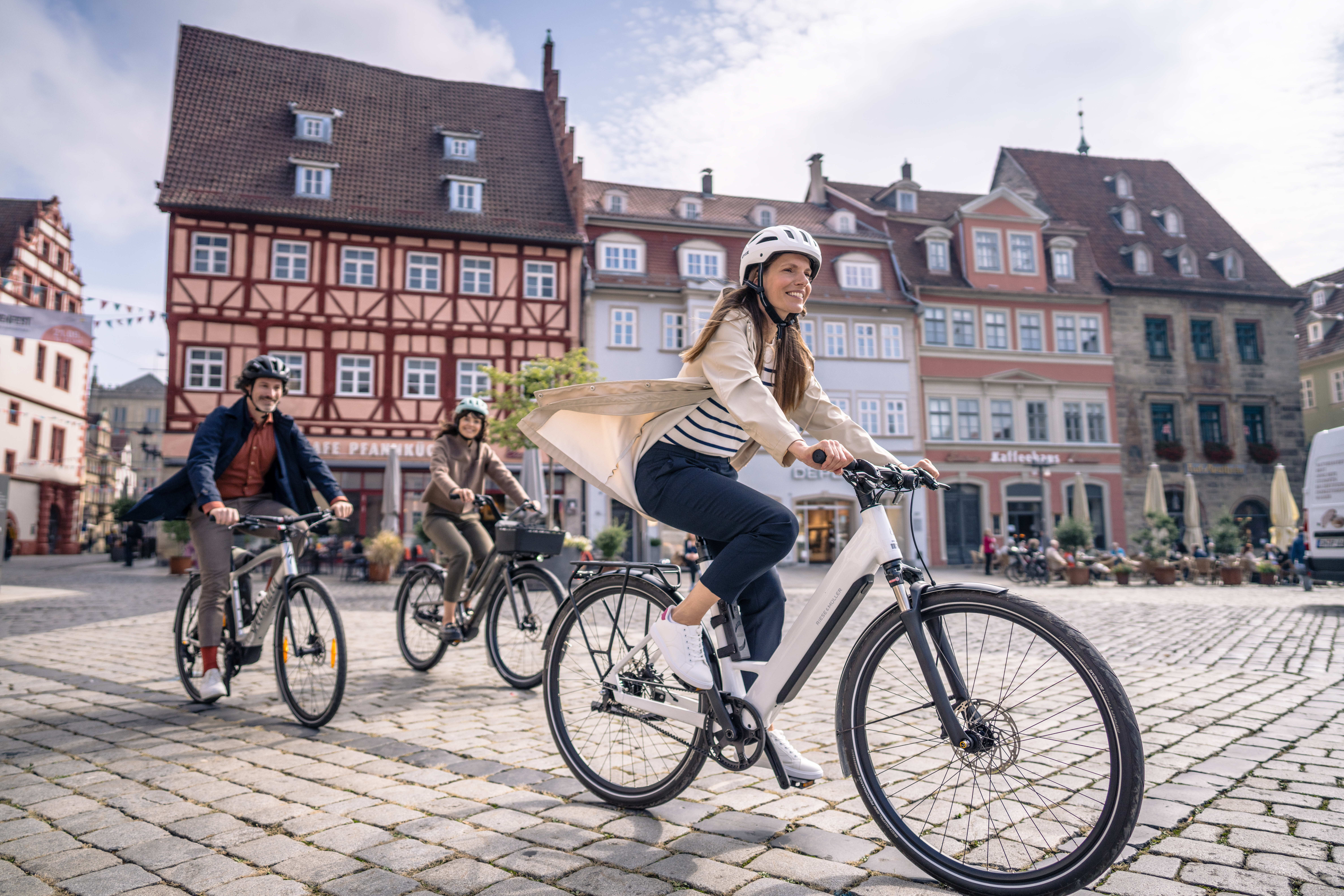 Drei fröhliche Menschen fahren mit E-Bikes über einen Kopfsteinpflasterplatz vor historischen Fachwerkhäusern.