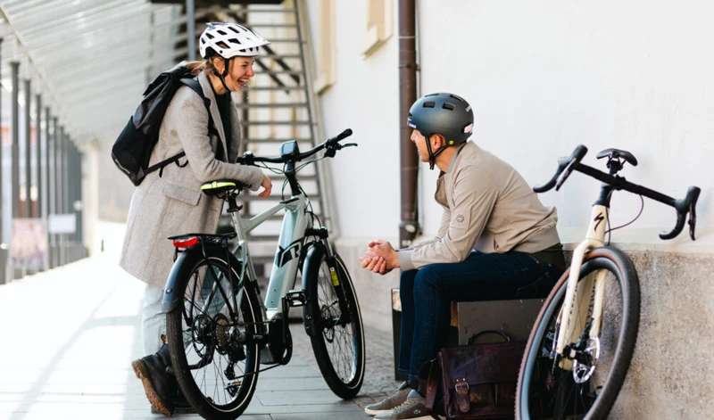 Eine Frau und ein Mann, beide mit Fahrradhelmen, unterhalten sich lächelnd. Die Frau steht neben einem E-Bike, während der Mann auf einer Bank sitzt, neben ihm ein Rennrad.