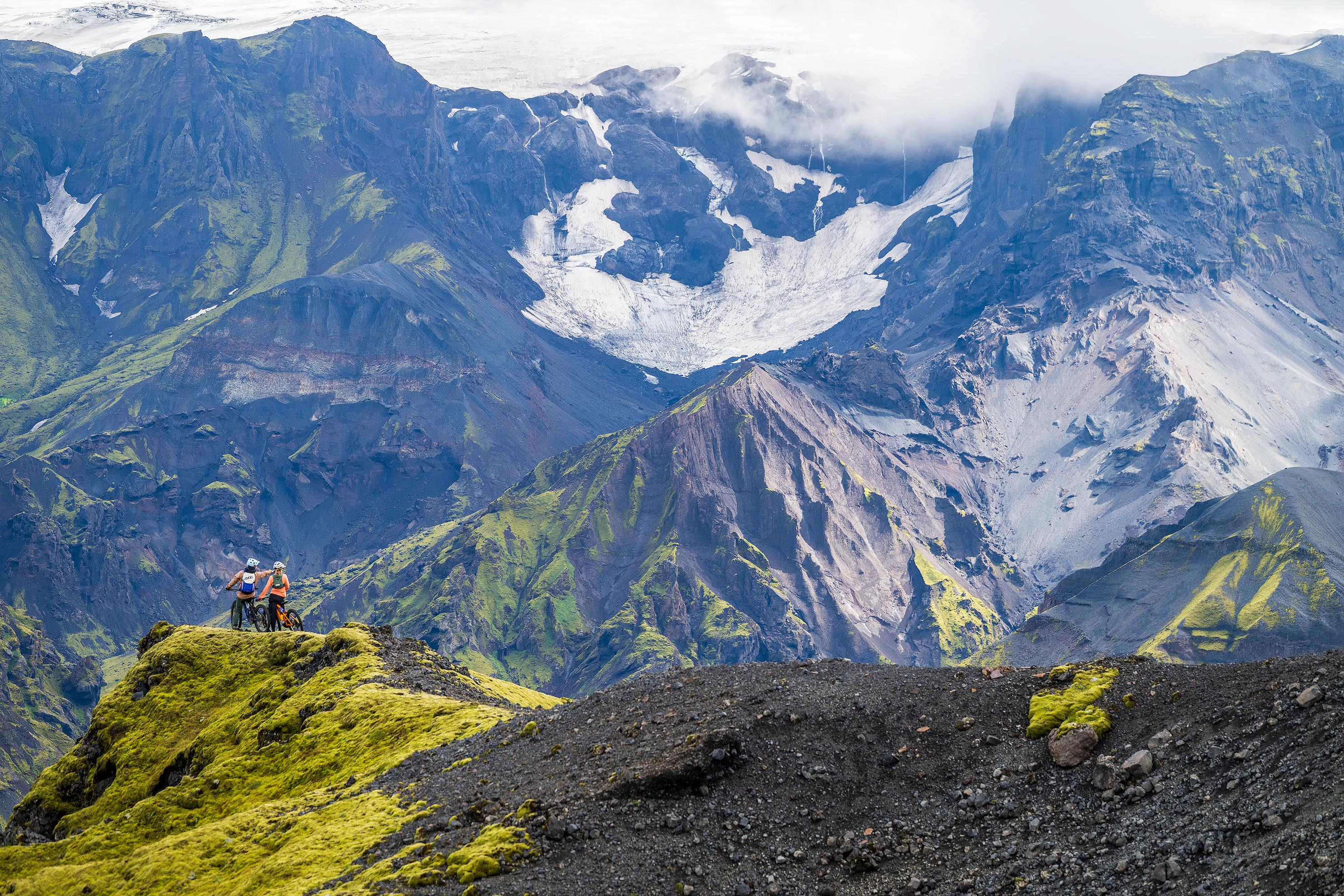 Zwei Mountainbiker stehen auf einem moosigen Grat und blicken auf eine dramatische Berg- und Gletscherlandschaft unter tiefhängenden Wolken.