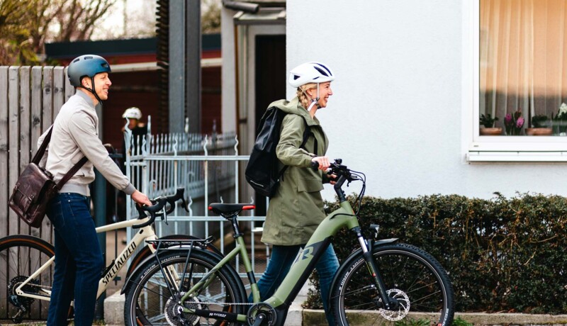 Ein Mann und eine Frau, beide mit Fahrradhelmen, schieben ihre Fahrräder entlang eines Gehwegs. Die Frau trägt einen Rucksack, und beide scheinen auf dem Weg zu sein.