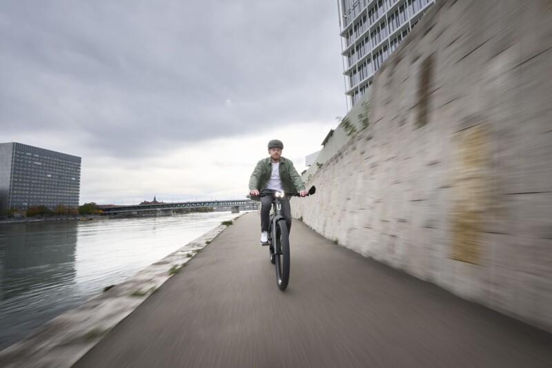 Ein Mann mit Helm fährt auf einem E-Bike vor einer Steinmauer, während der Himmel wolkenverhangen ist.