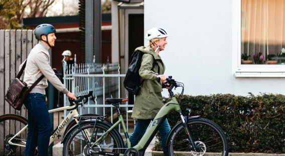 Ein Mann und eine Frau, beide mit Fahrradhelmen, schieben ihre Fahrräder entlang eines Gehwegs. Die Frau trägt einen Rucksack, und beide scheinen auf dem Weg zu sein.