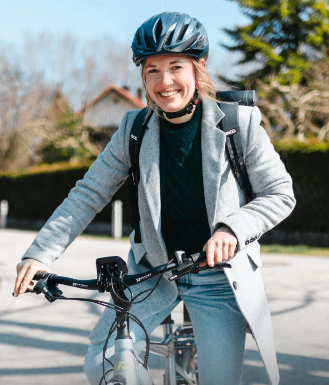 Eine Frau auf einem Fahrrad trägt einen Fahrradhelm und lächelt in die Kamera. Sie ist in einen grauen Mantel und eine Jeans gekleidet, der Hintergrund zeigt eine Straße und Bäume.