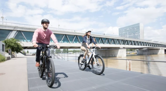 Eine Frau und ein Mann, beide mit Fahrradhelmen, unterhalten sich lächelnd. Die Frau steht neben einem E-Bike, während der Mann auf einer Bank sitzt, neben ihm ein Rennrad.