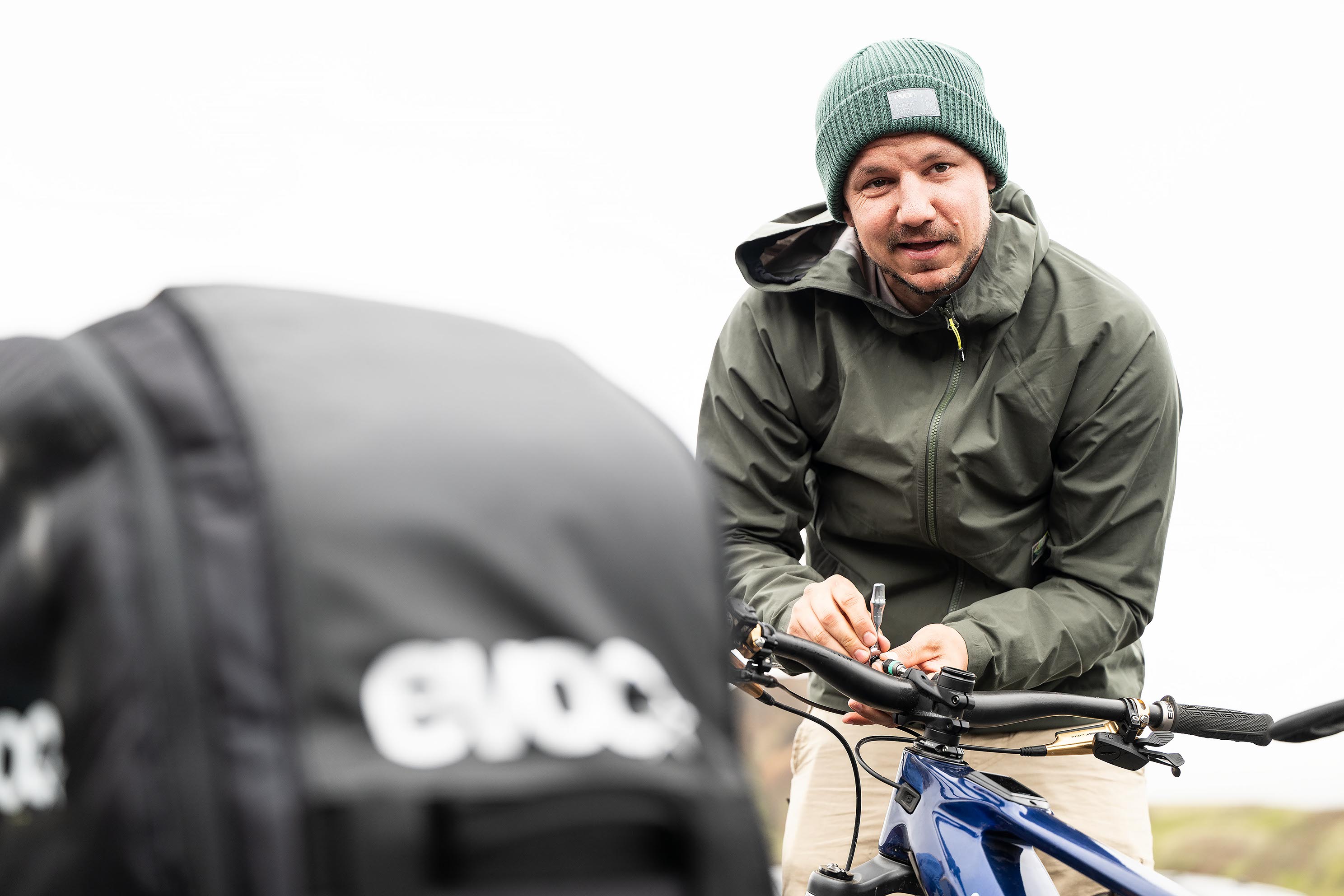 Ein Mann mit grüner Mütze und Regenjacke beugt sich über den Lenker eines Fahrrads und stellt etwas ein, während im Vordergrund eine unscharfe Tasche zu sehen ist.