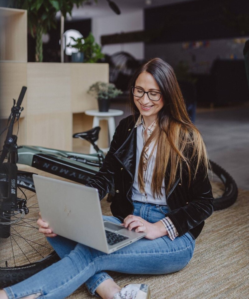 Eine Frau sitzt auf dem Boden neben einem Fahrrad und arbeitet lächelnd an ihrem Laptop. Im Hintergrund sind Pflanzen und ein moderner, offener Raum zu sehen.