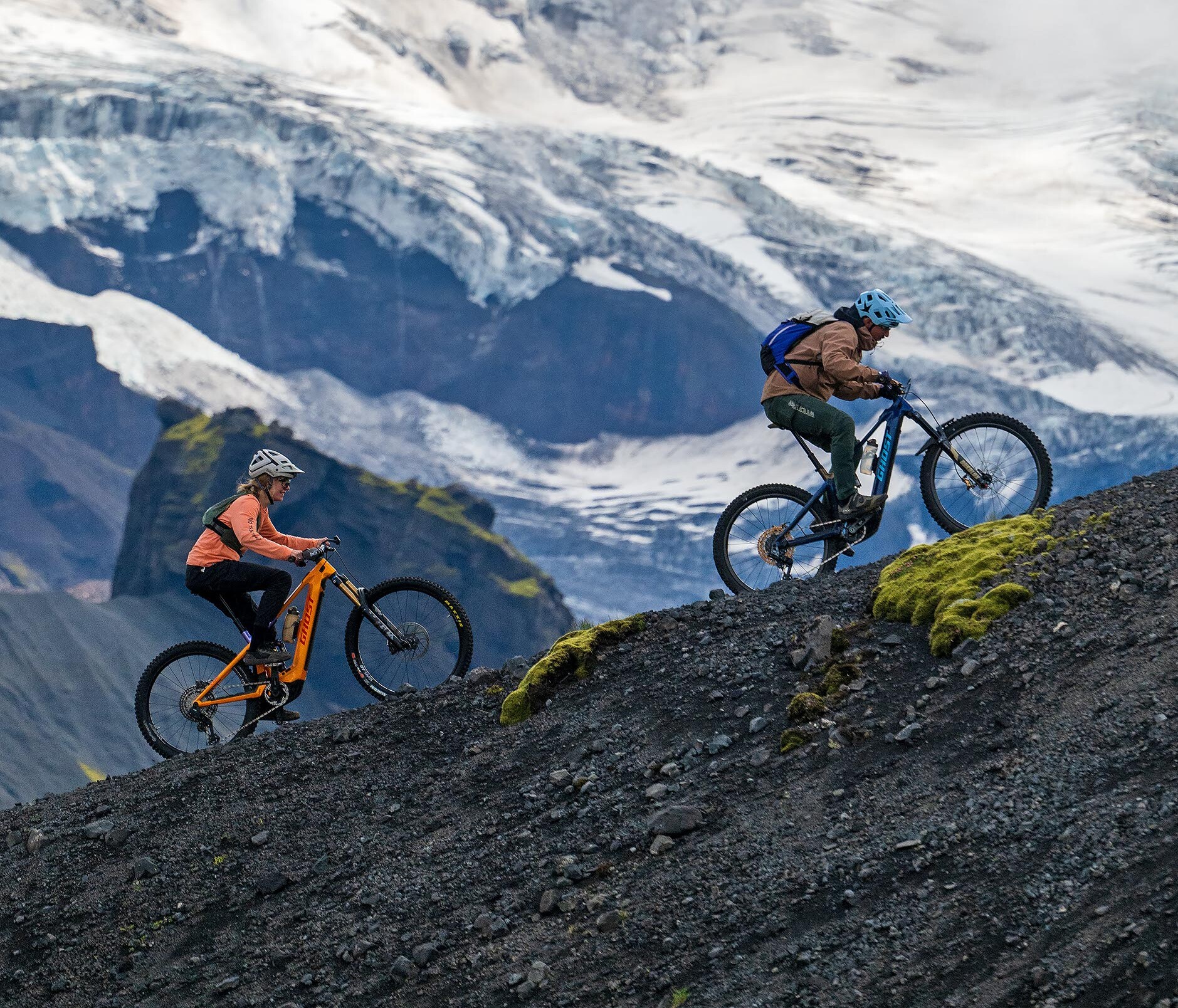 Zwei Mountainbiker mit Helmen fahren einen steilen, schwarzen Geröllhang hinauf, vor einer dramatischen Gletscher- und Bergkulisse.
